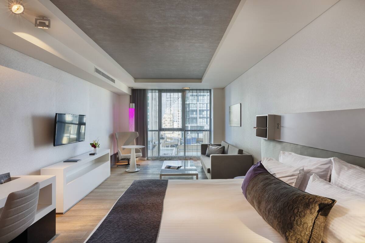 A modern hotel room with a large bed, a flat-screen TV, a sofa, a coffee table, and a view of the city through large windows.