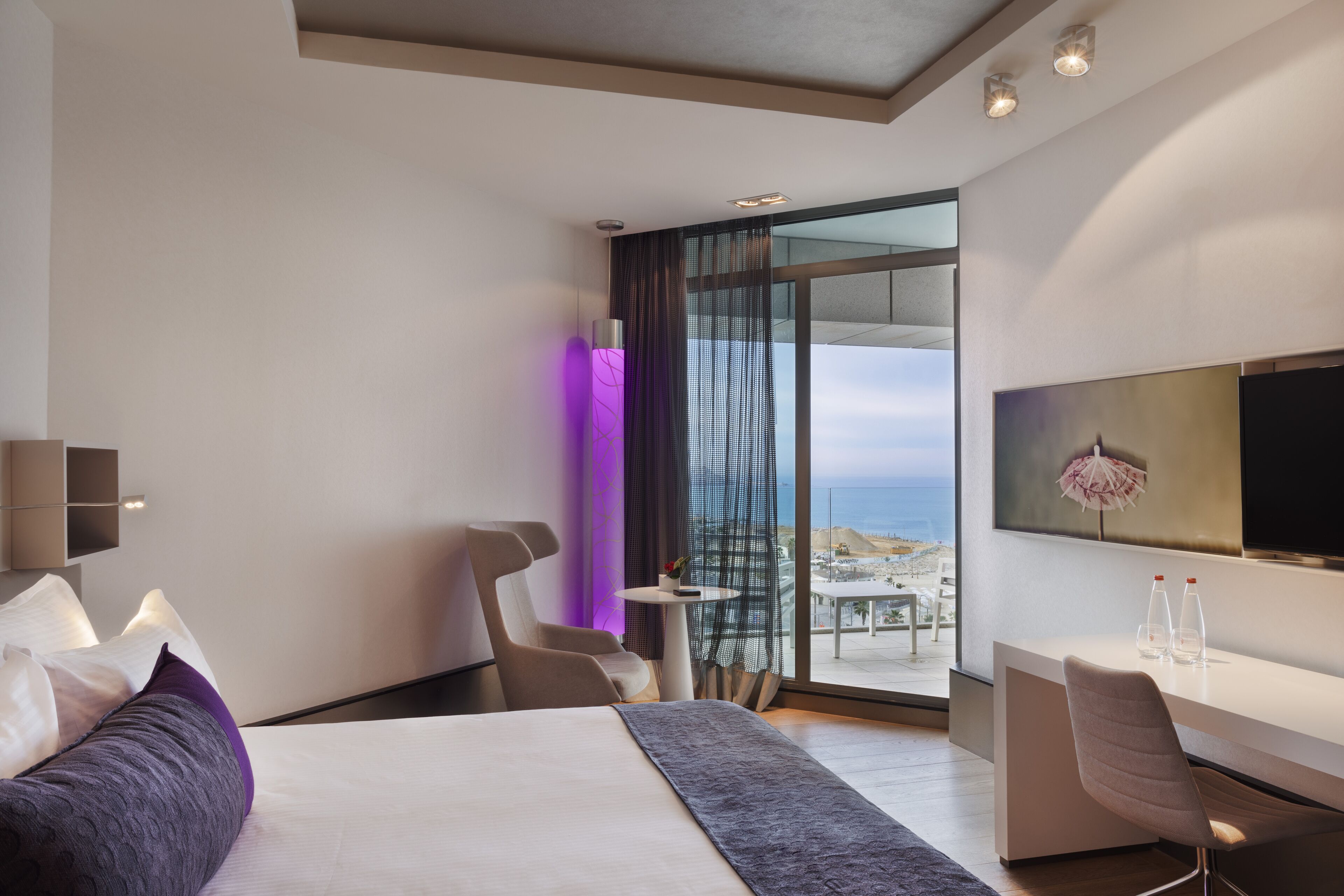 Une chambre d’hôtel moderne équipée d’un lit, d’un bureau et d’une grande fenêtre donnant sur la plage.