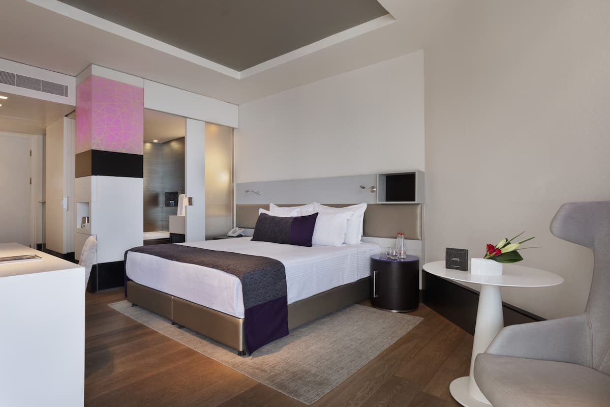 A modern hotel room with a large bed, bedside tables, a small round table with a vase, and a chair.