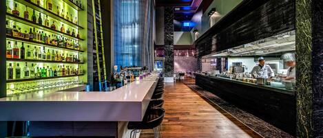 A bar area with a long counter, multiple shelves of bottles, and a kitchen in the background.
