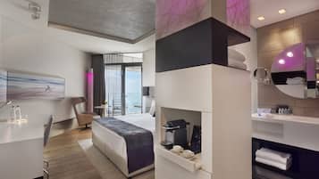 Quarto de hotel moderno com cama, escrivaninha, cadeira, banheiro com espelho e vista para o mar.