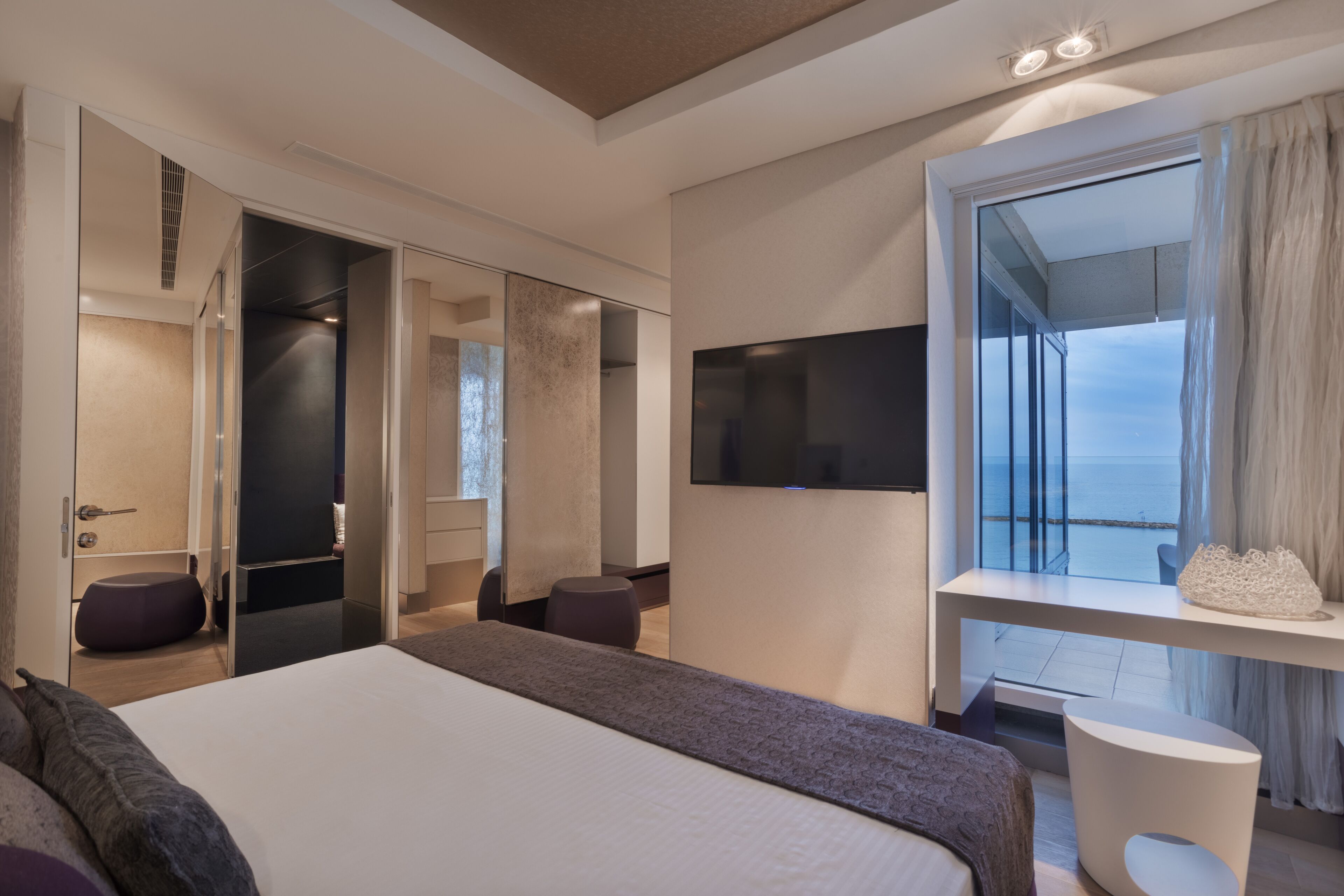 A modern hotel room with a large bed, a TV mounted on the wall, and a balcony with a view of the sea.