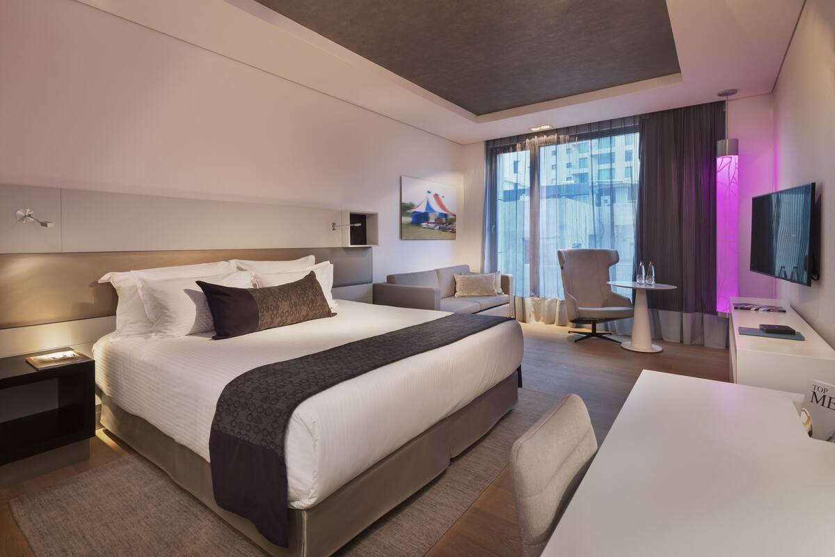 A modern hotel room with a large bed, a desk, a chair, a TV, and a window with curtains.