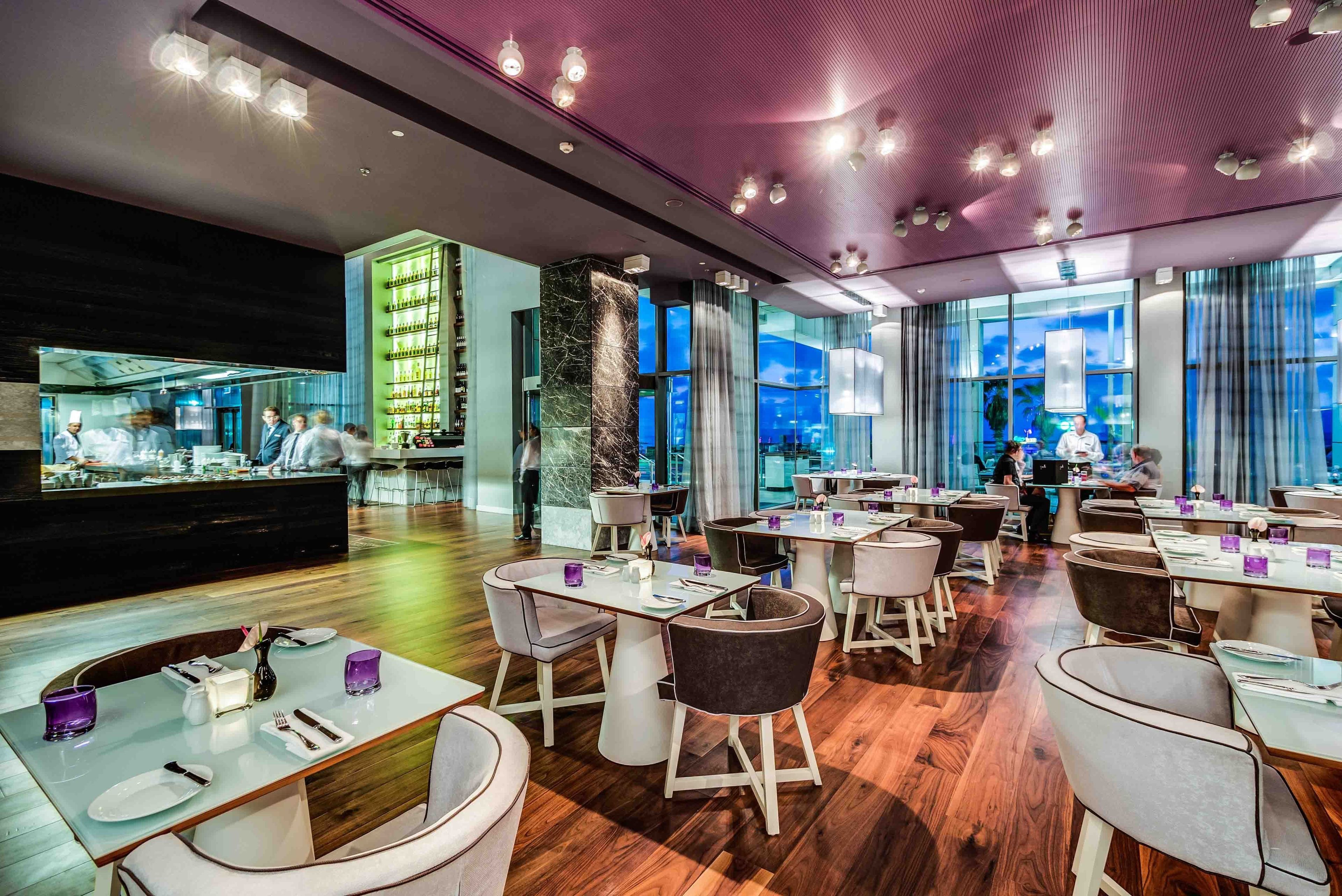 A modern restaurant with a central bar area, multiple dining tables, and a view of the cityscape through large windows.