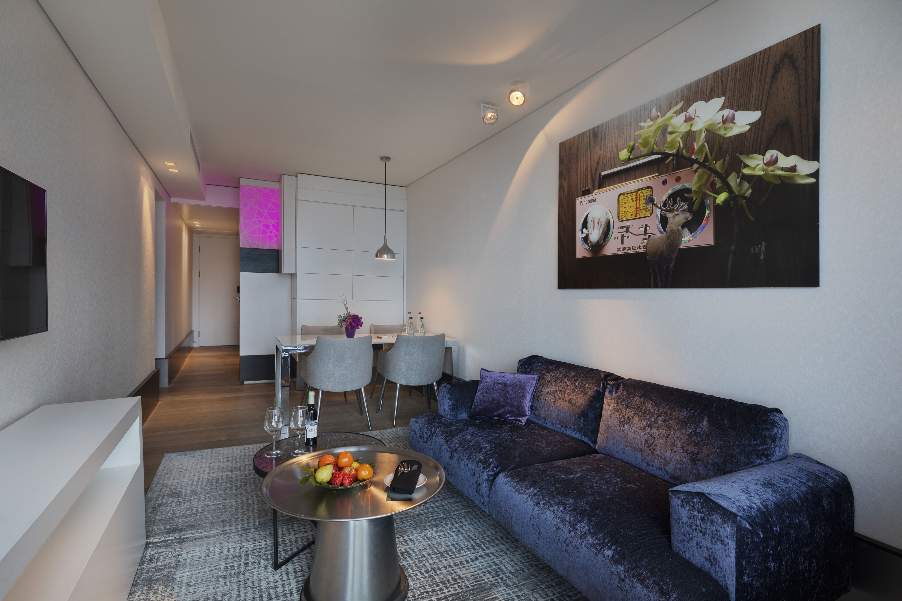 Un salon moderne avec un canapé bleu foncé, une table basse ronde sur laquelle sont disposés des fruits, et un coin repas à l’arrière-plan.