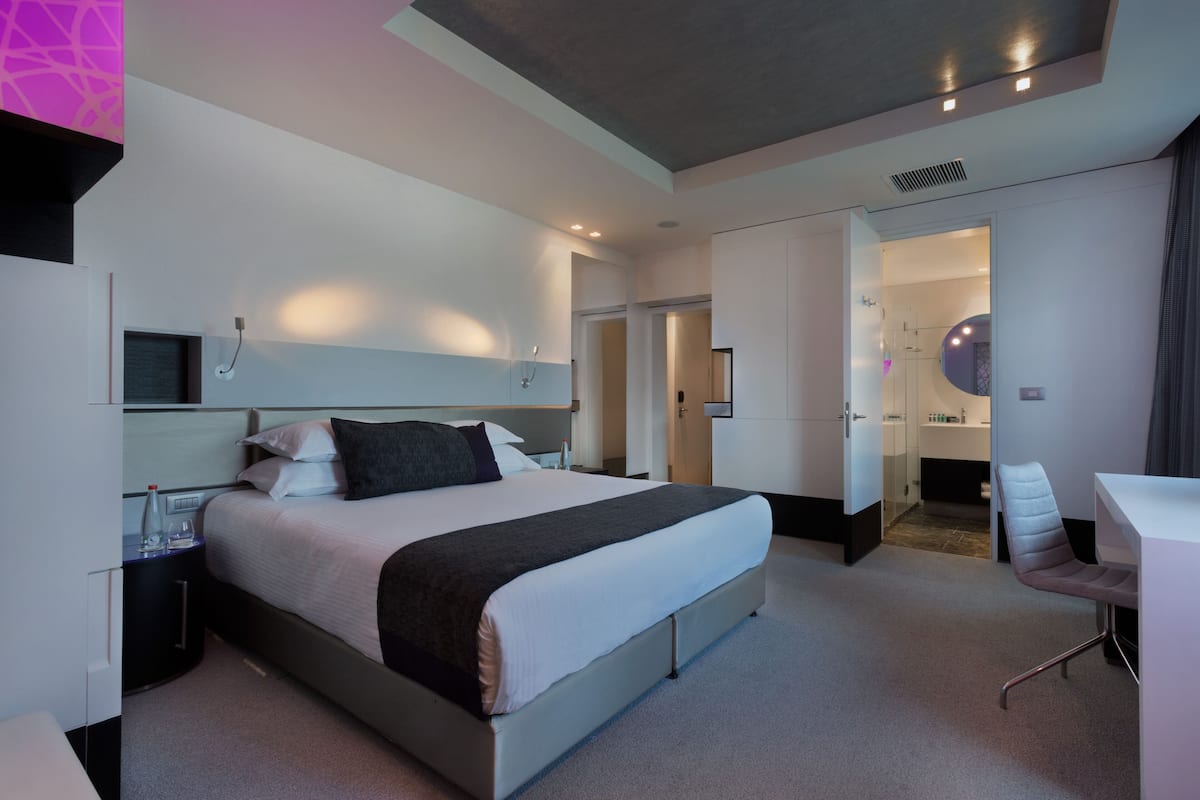 A modern hotel room with a large bed, bedside tables, a desk, and a bathroom visible through an open door.