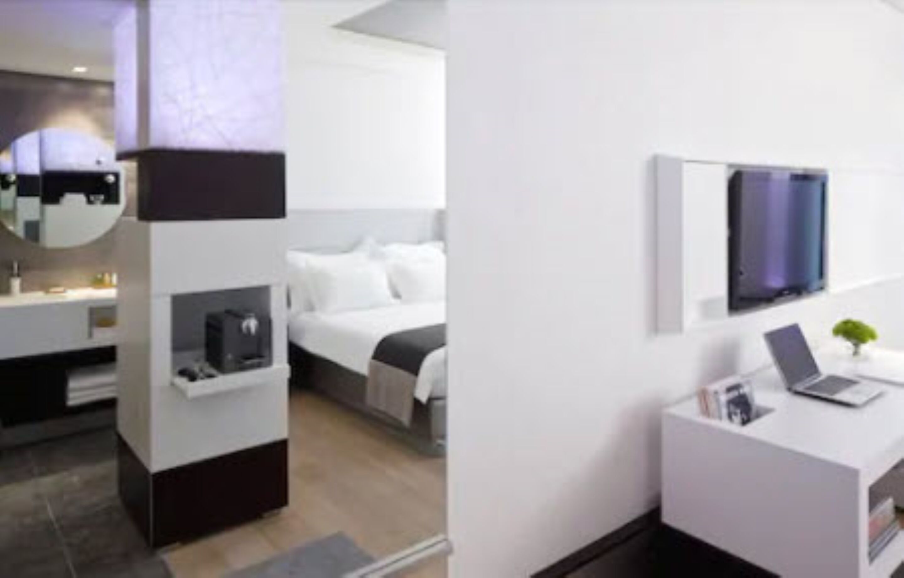 A modern hotel room with a bed, a desk with a laptop, a TV, and a small plant.