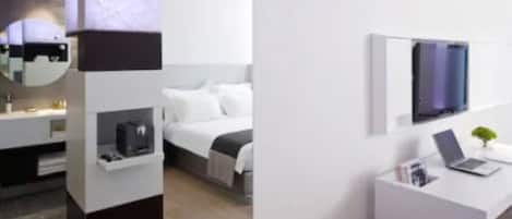 A modern hotel room with a bed, a desk with a laptop, a TV, and a small plant.