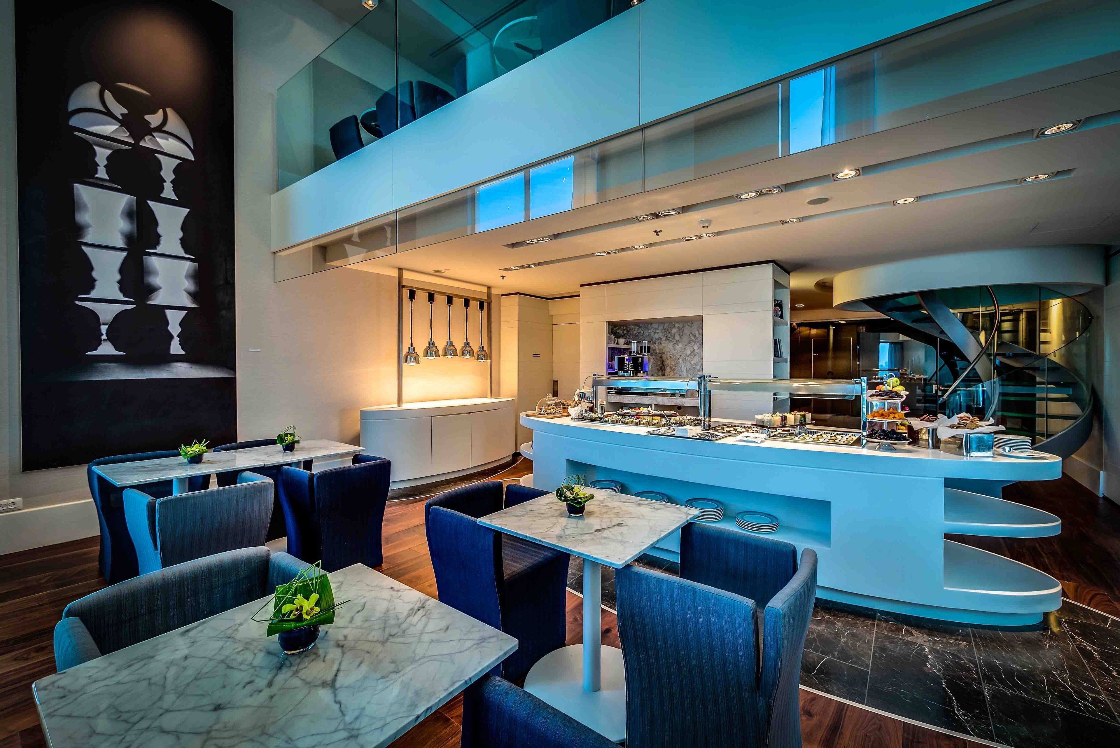 A modern restaurant with marble tables and blue chairs, a central bar area with food and drinks, and a staircase leading to an upper level.