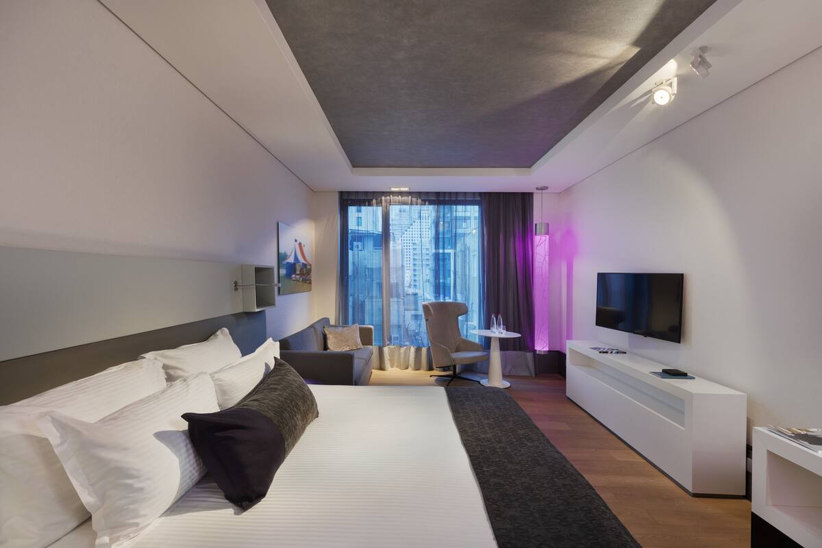 A modern hotel room with a large bed, a TV, a desk, and a chair.