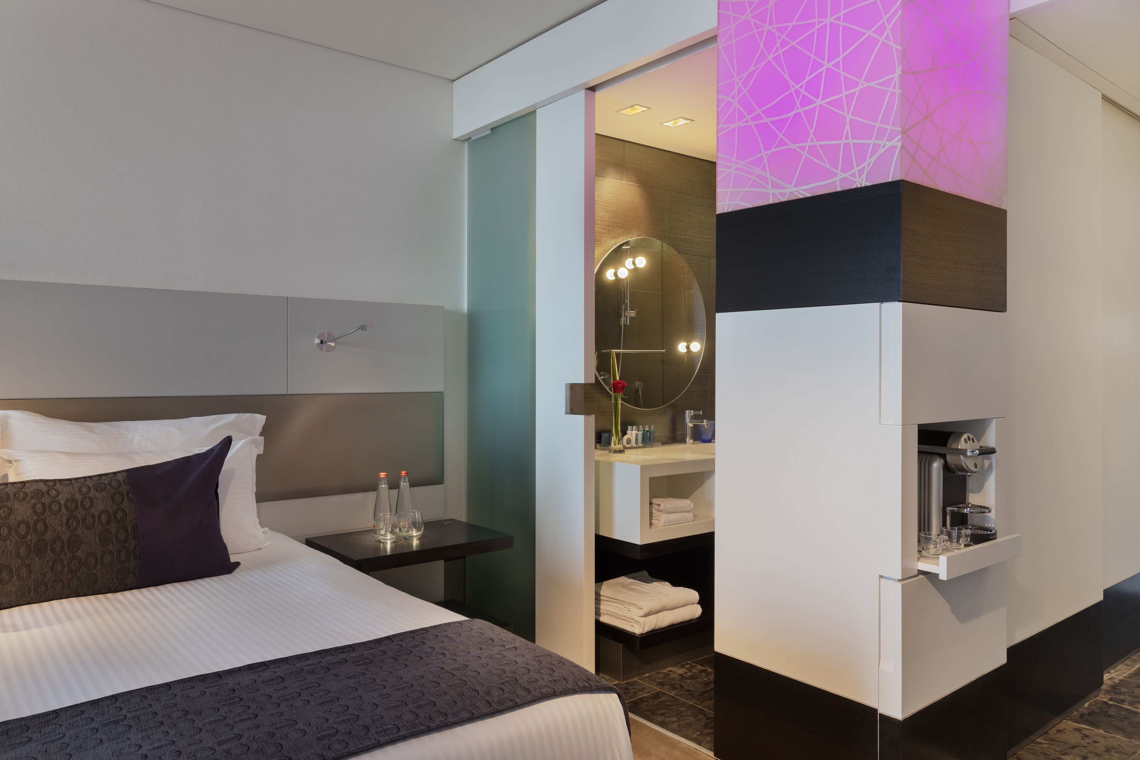 Una habitación de hotel moderna con una cama bien hecha, una mesita de noche con lámpara, un tocador con espejo y una cafetera integrada.
