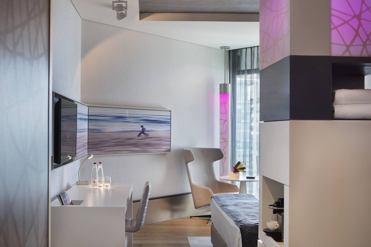 A modern hotel room with a bed, a desk with a computer, a chair, a small table with a lamp, and a window with a view of the city.