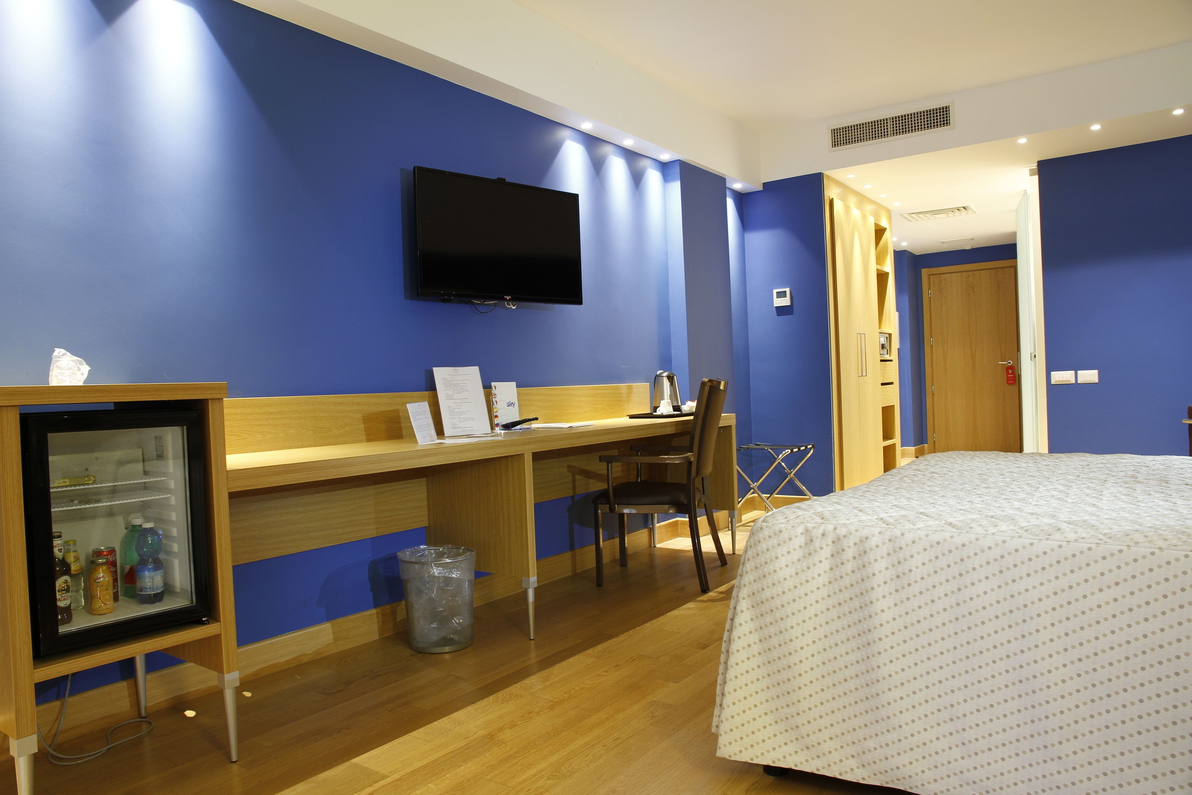 A hotel room with a bed, a desk with a chair, a television mounted on the wall, and a small refrigerator.
