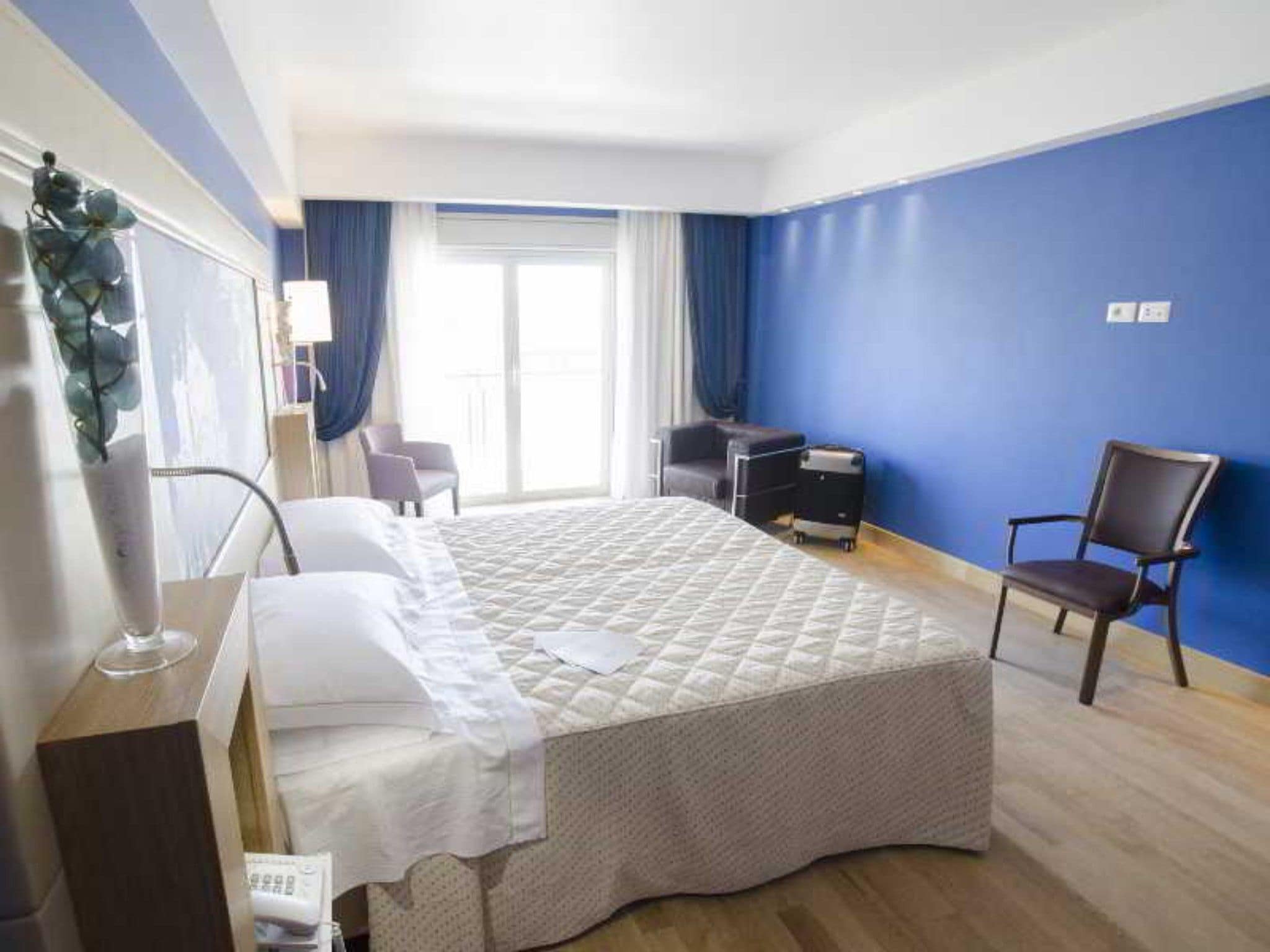 A hotel room with a bed, a desk, a chair, a desk lamp, and a window with curtains.