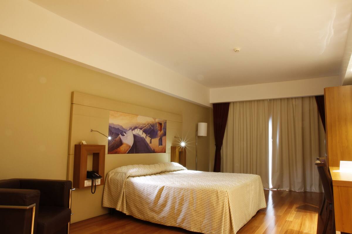 Una habitación de hotel con cama, escritorio, silla, teléfono, un cuadro en la pared y ventana con cortinas.