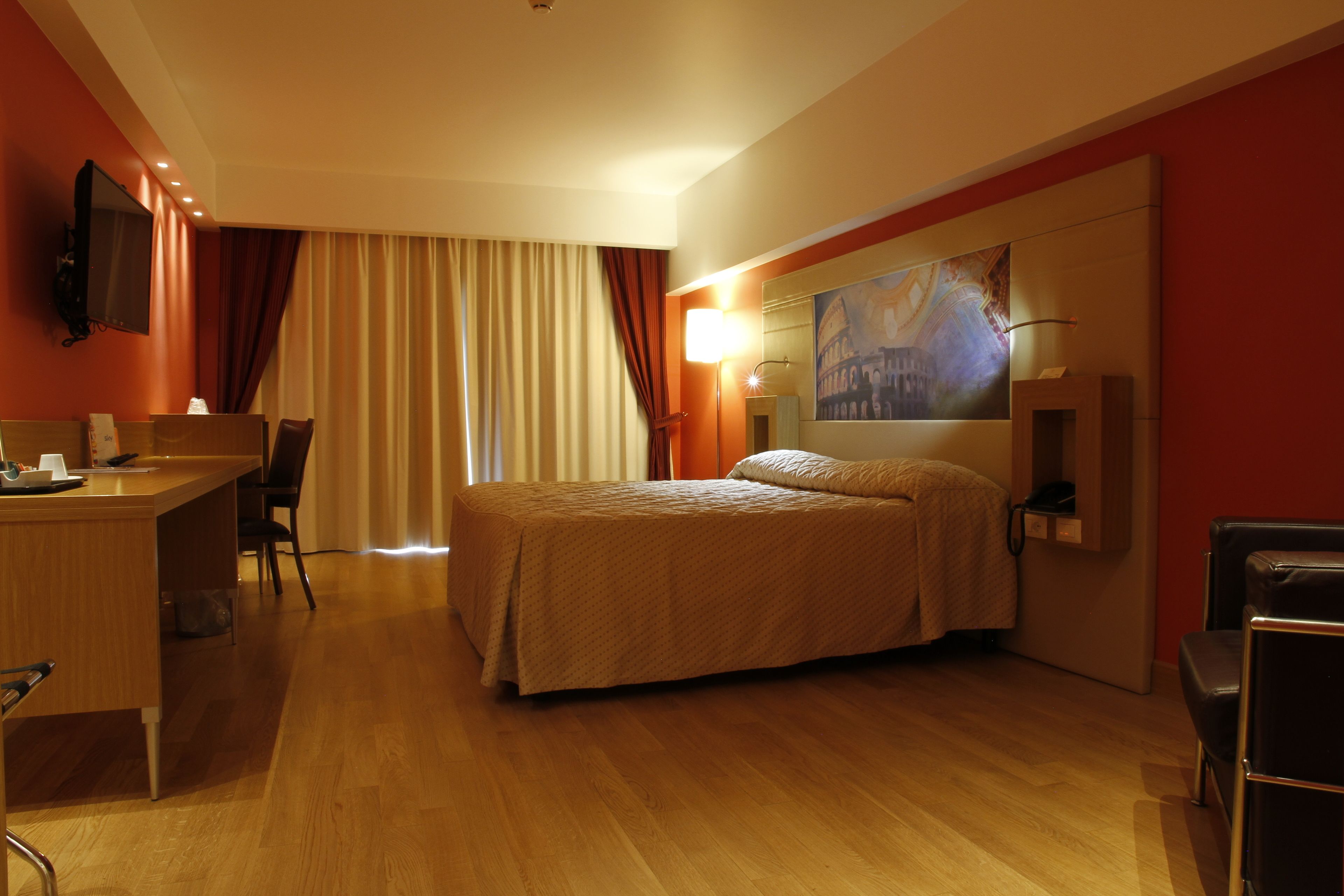 A hotel room with a bed, desk, chair, and a painting on the wall.
