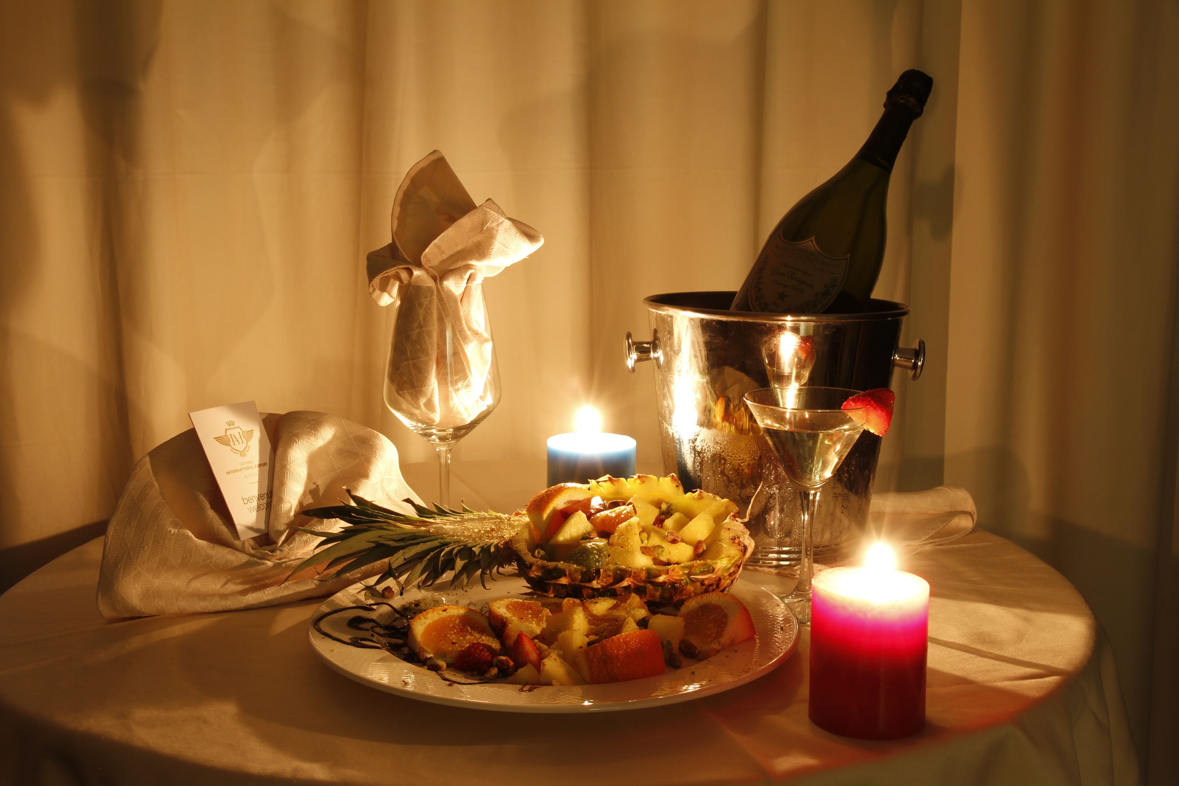 A table with a bottle of champagne, a candle, a glass, and a plate of fruit.