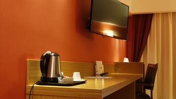 A hotel room with a wooden desk, a television mounted on the wall, a coffee maker, and a chair.