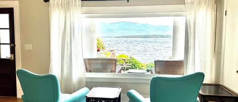 Una habitación con dos sillones turquesa, una mesita y vistas a una masa de agua a través de una ventana con cortinas blancas.