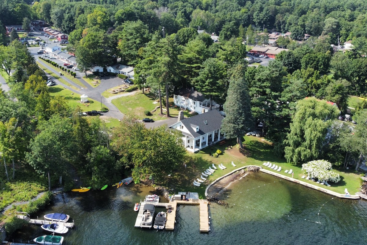 An aerial view of a lakeside property with a dock, boats, and a large house surrounded by dense trees.