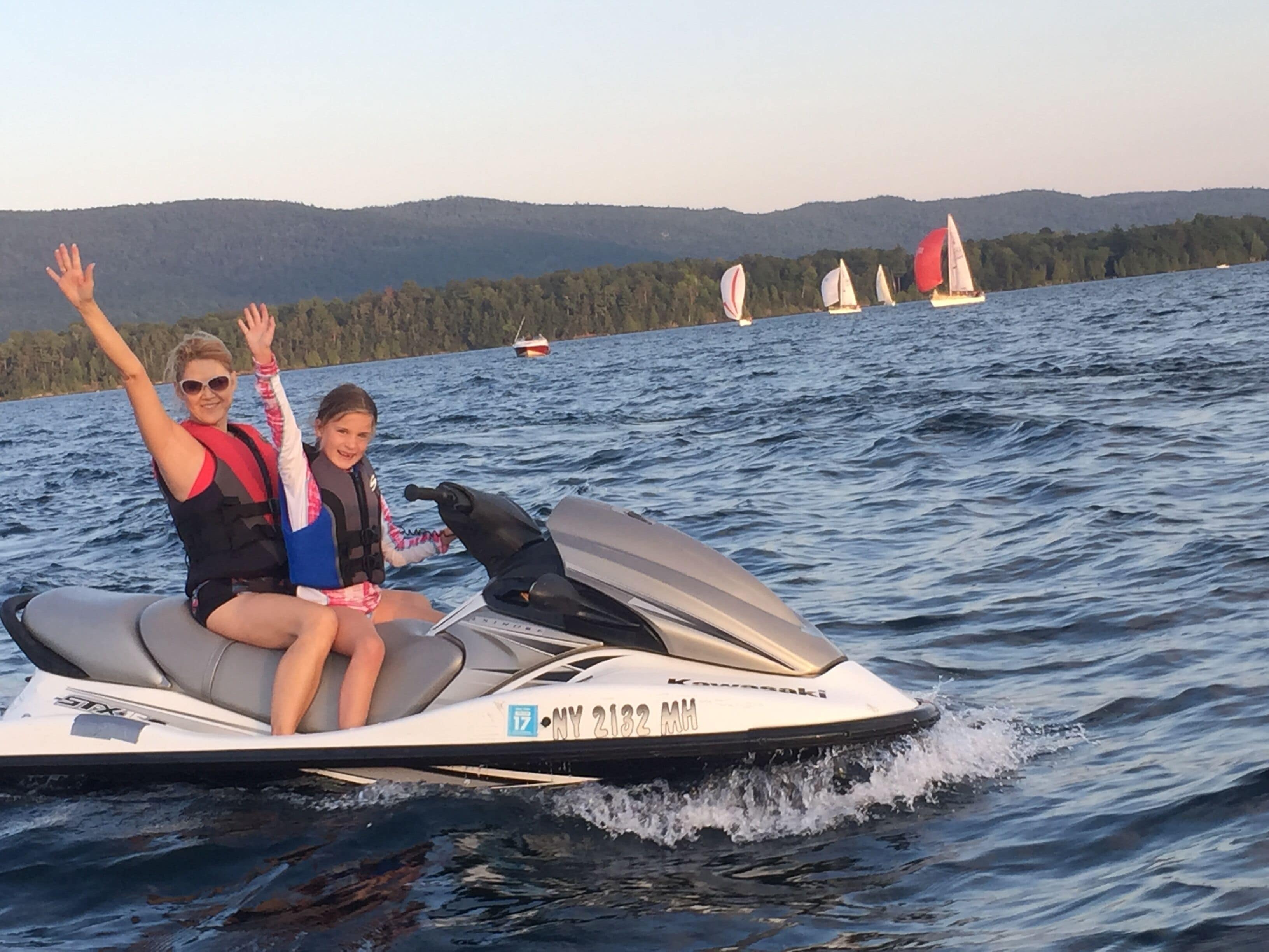 Deux personnes sur un jet ski, avec un lac et des voiliers en arrière-plan.