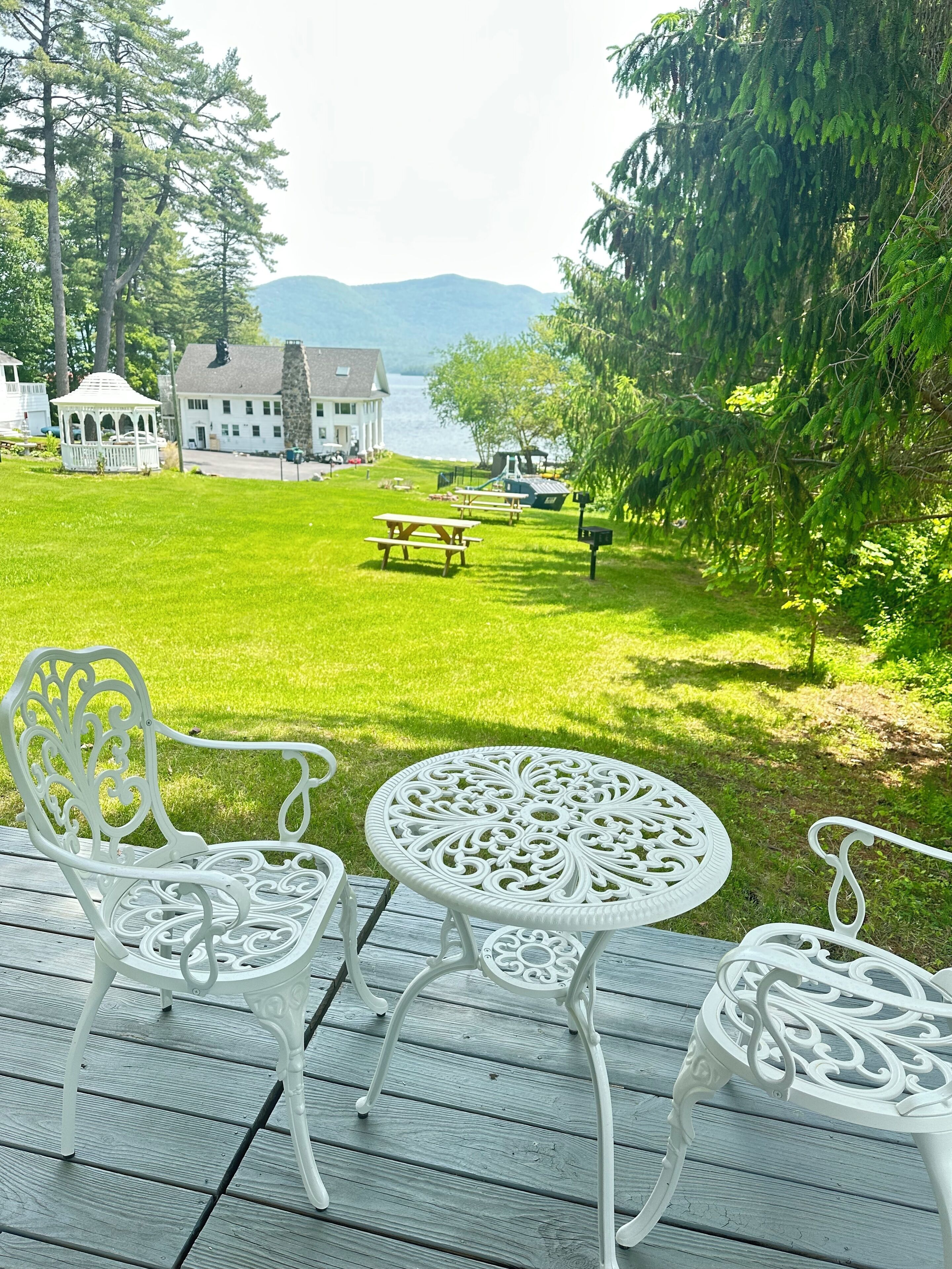 Une terrasse avec des chaises et une table en métal peintes en blanc, donnant sur une pelouse où se trouvent des tables de pique-nique et un kiosque.