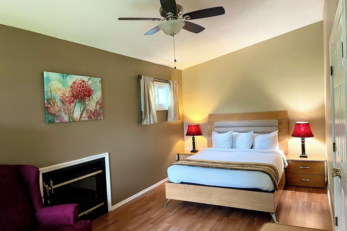 A bedroom with a bed, two bedside lamps, a ceiling fan, a fireplace, and a floral painting on the wall.