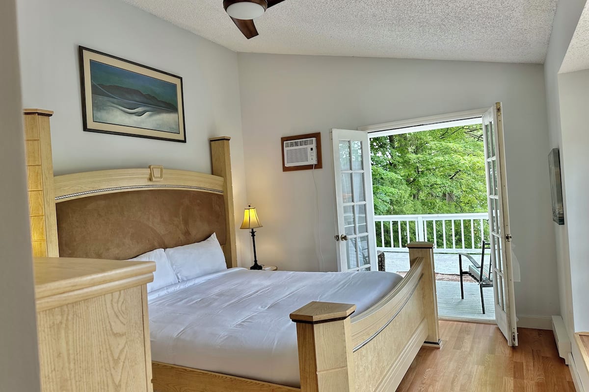 A bedroom with a wooden bed, a ceiling fan, a nightstand with a lamp, and a door leading to a balcony with a view of trees.