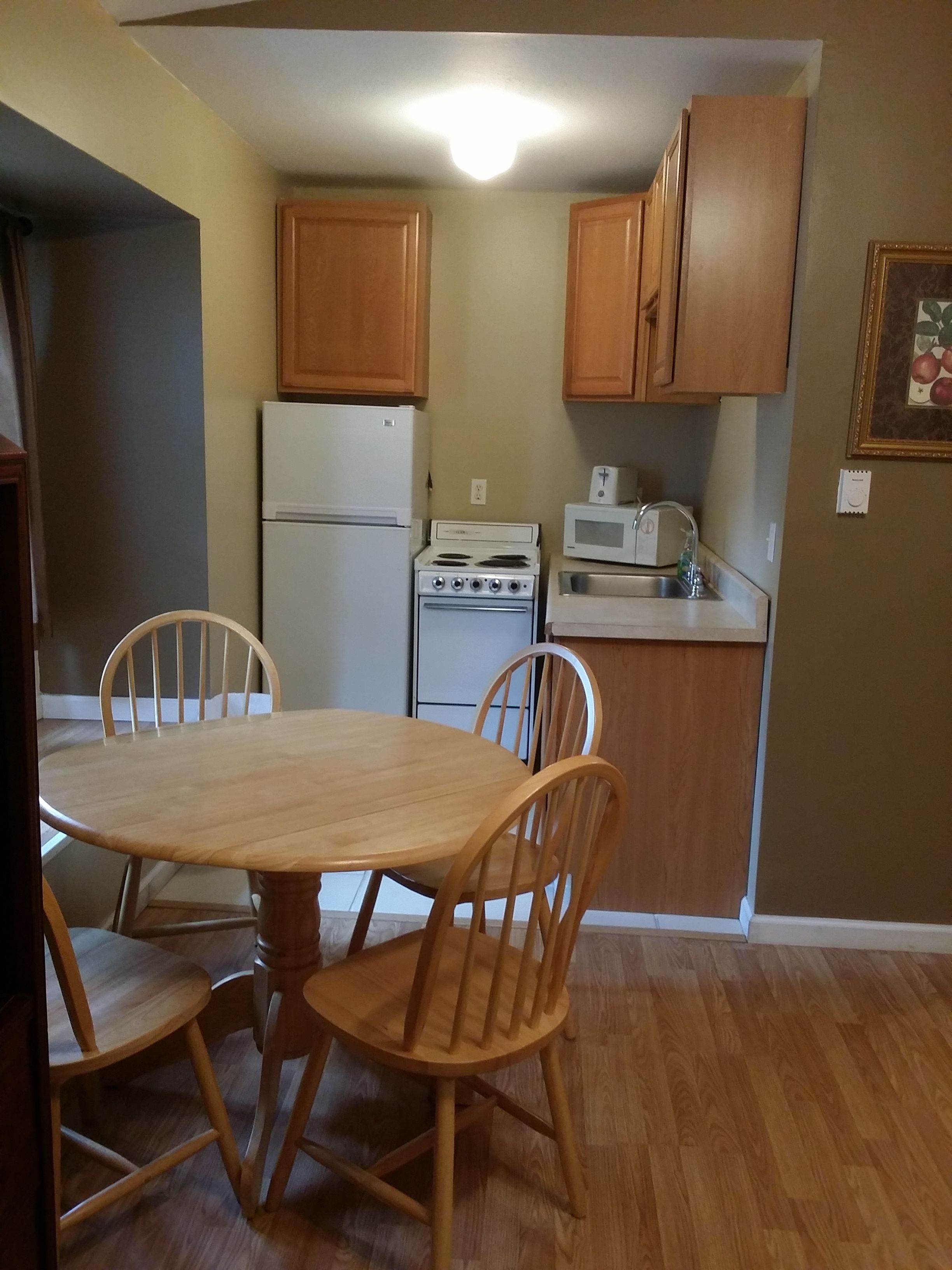 A compact kitchen with wooden cabinets, a refrigerator, a stove, and a microwave. There is a dining table with chairs in the foreground.
