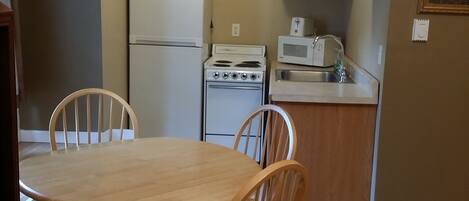 A compact kitchen with wooden cabinets, a refrigerator, a stove, and a microwave. There is a dining table with chairs in the foreground.