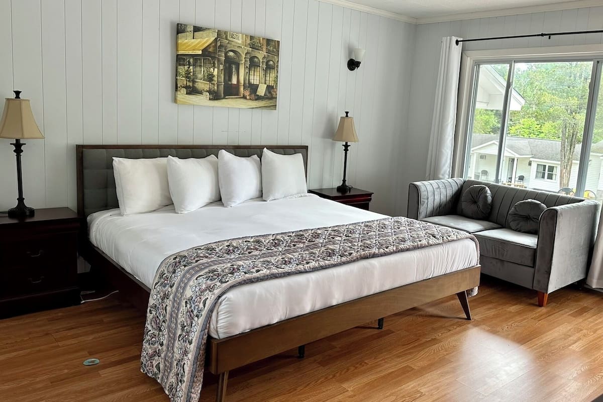 A bedroom with a large bed, two bedside lamps, a window with a view, and a painting on the wall.