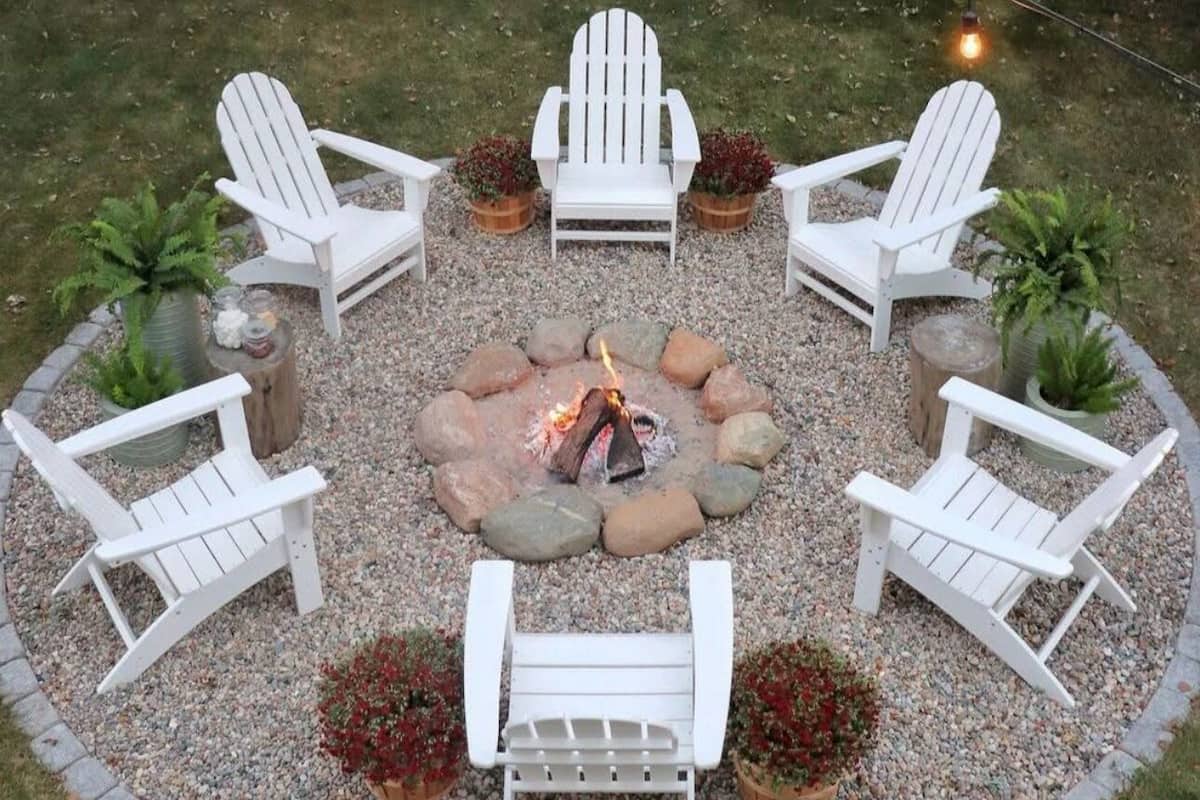 A circular outdoor seating area with Adirondack chairs around a central fire pit, surrounded by gravel and potted plants.