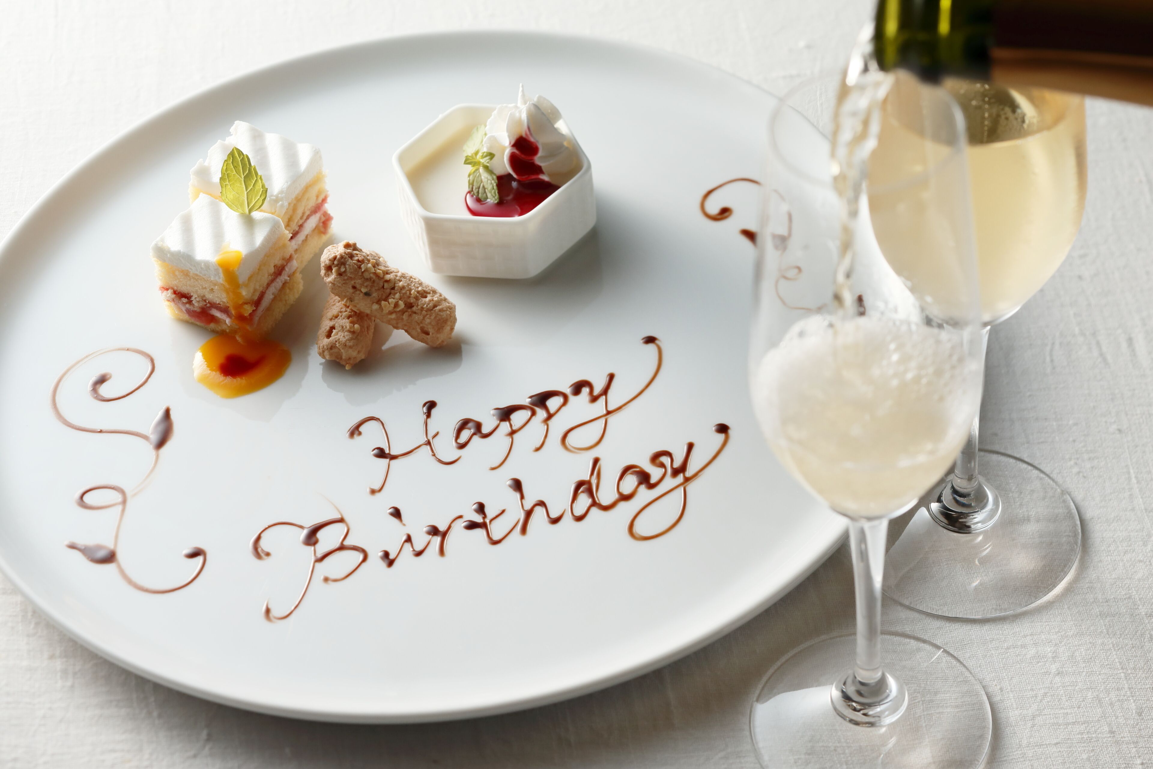 Ein weißer Teller mit einer Dessertanrichtung und eine Champagnerflasche, aus der Sekt in ein Glas gegossen wird.
