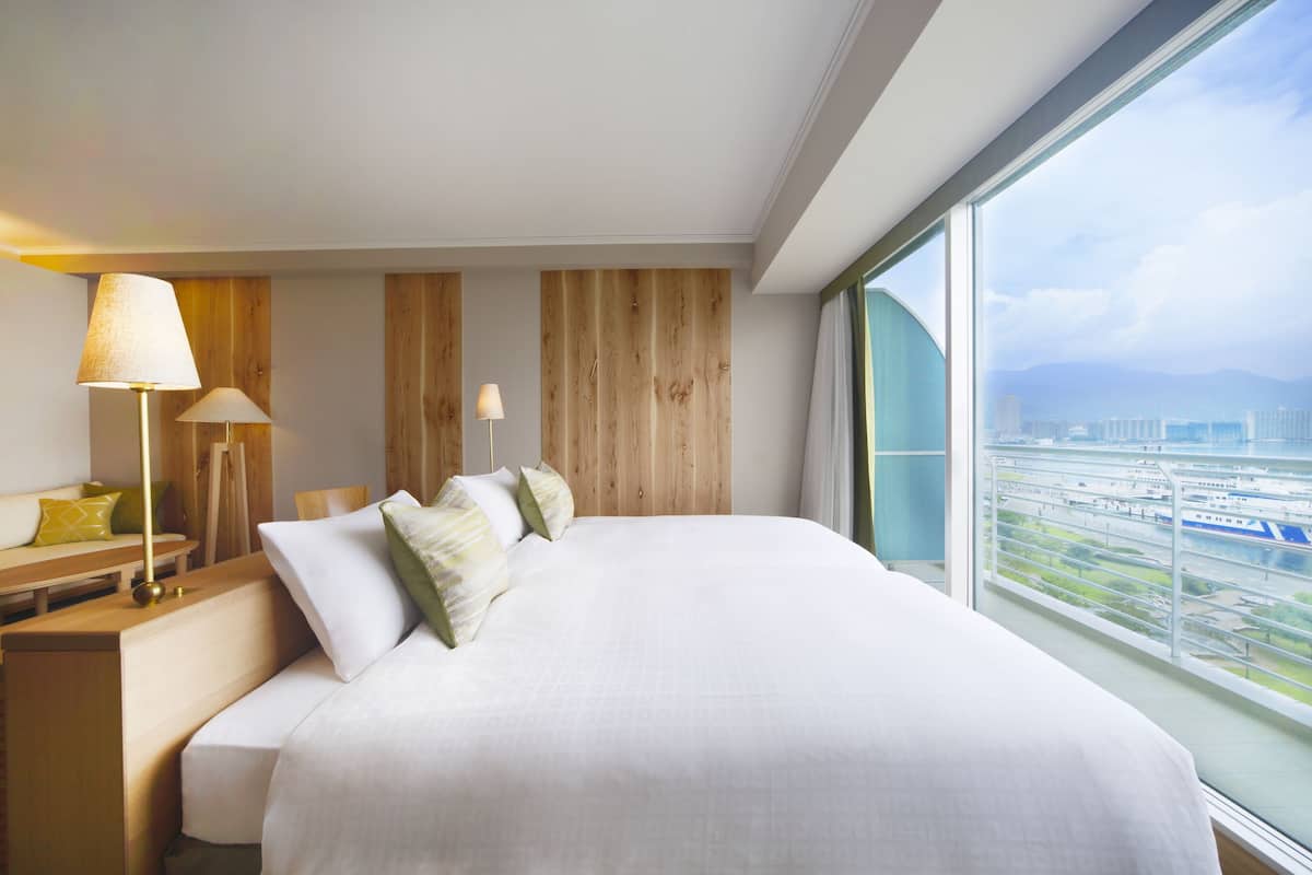 A modern hotel room with a large bed, wooden panel wall, and a view of a cityscape through a large window.