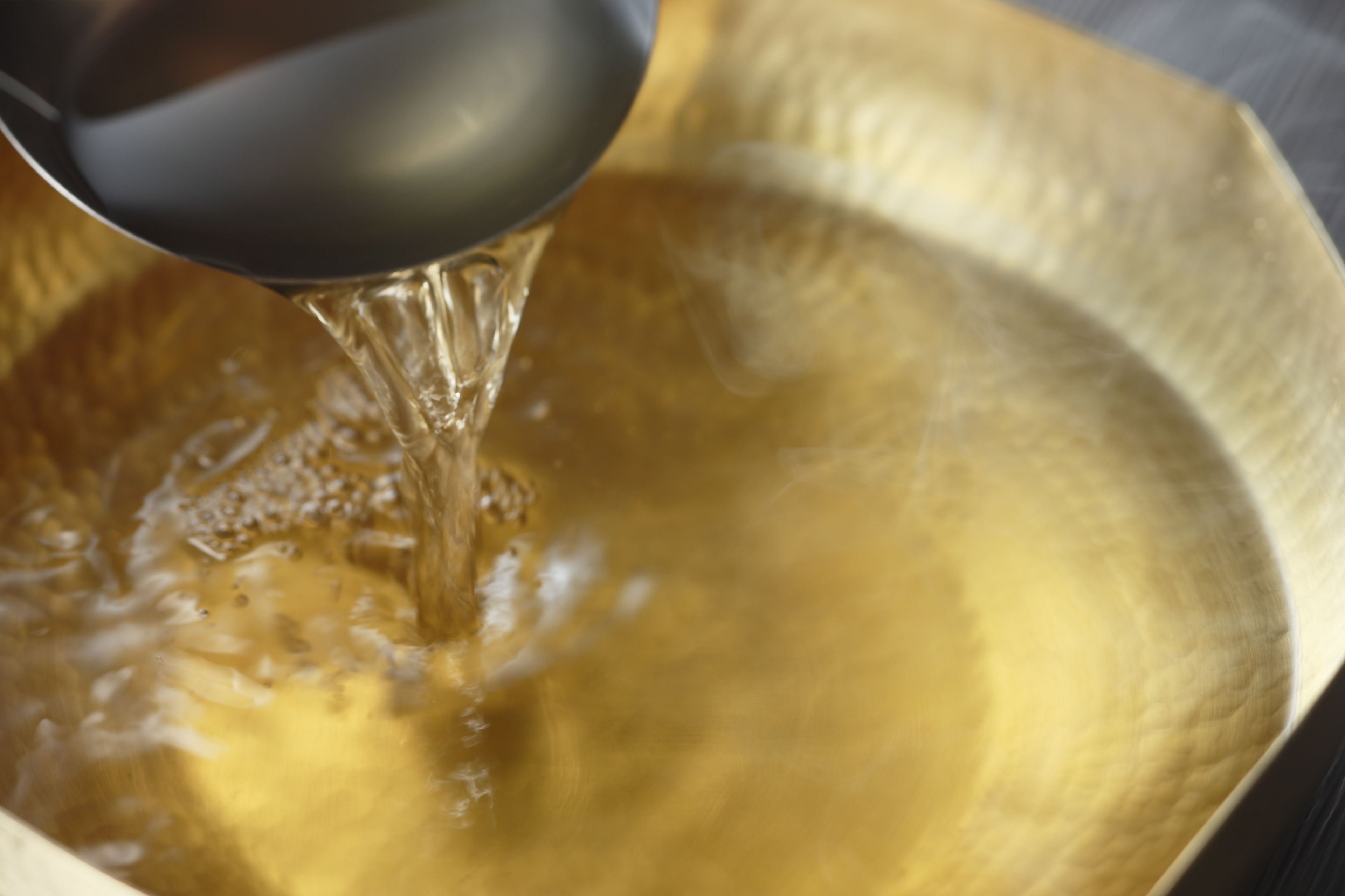 Un gros plan d’une cuillère métallique versant un liquide transparent dans un bol à surface texturée.