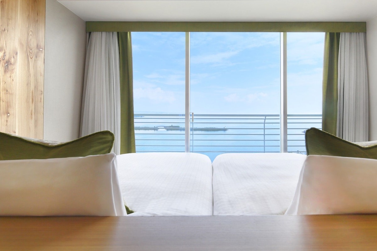 Een hotelkamer met een groot raam dat uitzicht biedt op de zee en een pier. Er staan twee witte bedden met groene kussens.