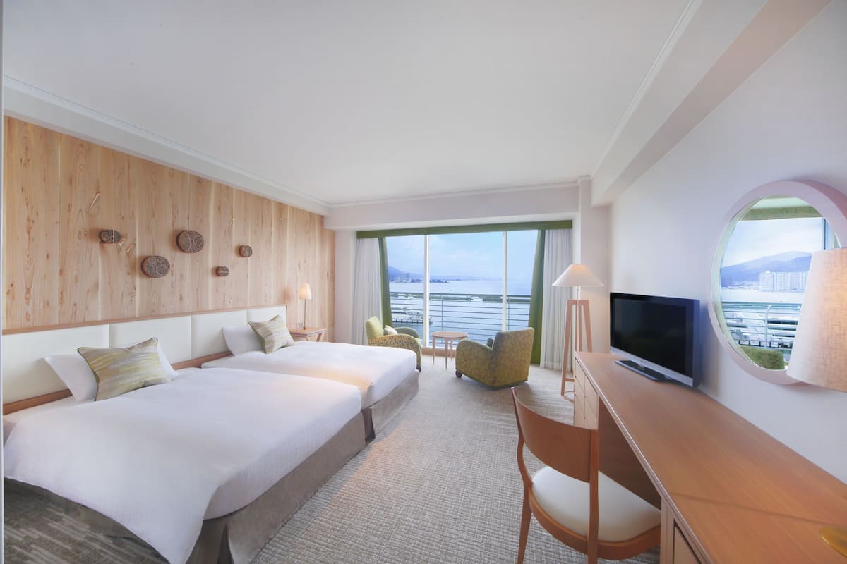 A hotel room with two beds, a desk with a TV, a chair, a lamp, and a large window with a view of the sea.