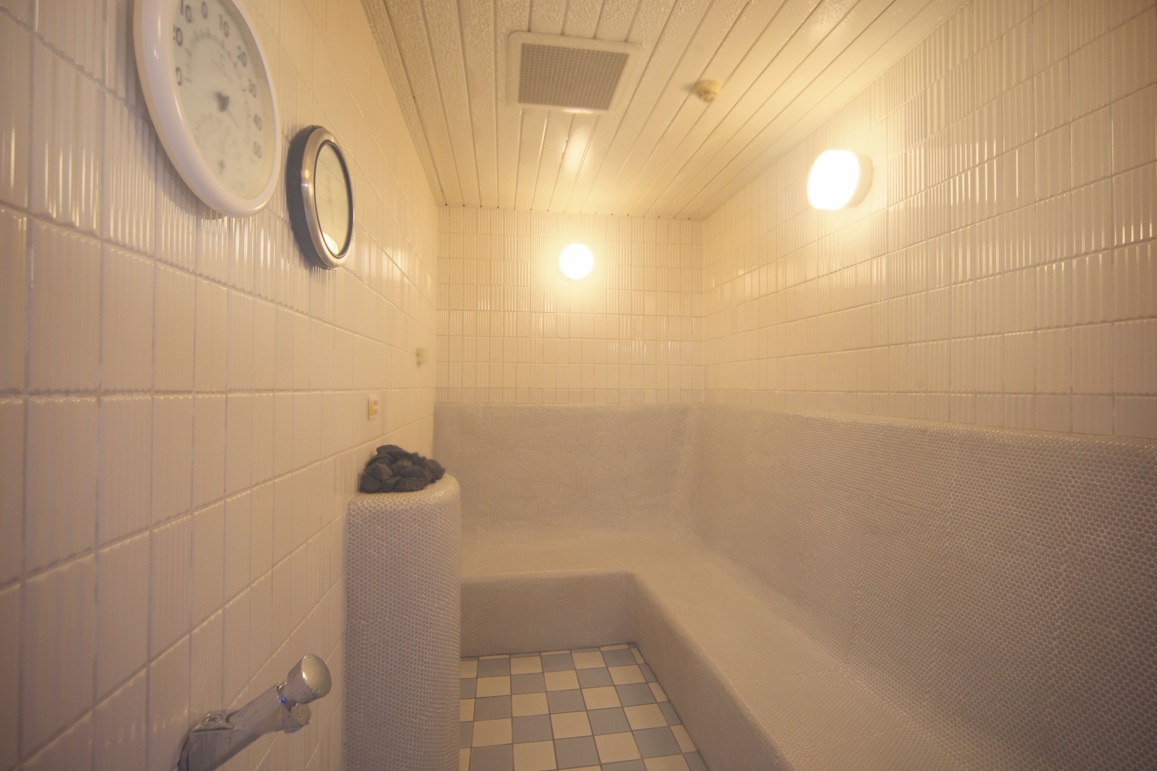 Un baño con paredes de azulejos blancos, piso a cuadros, una bañera y dos relojes de pared.