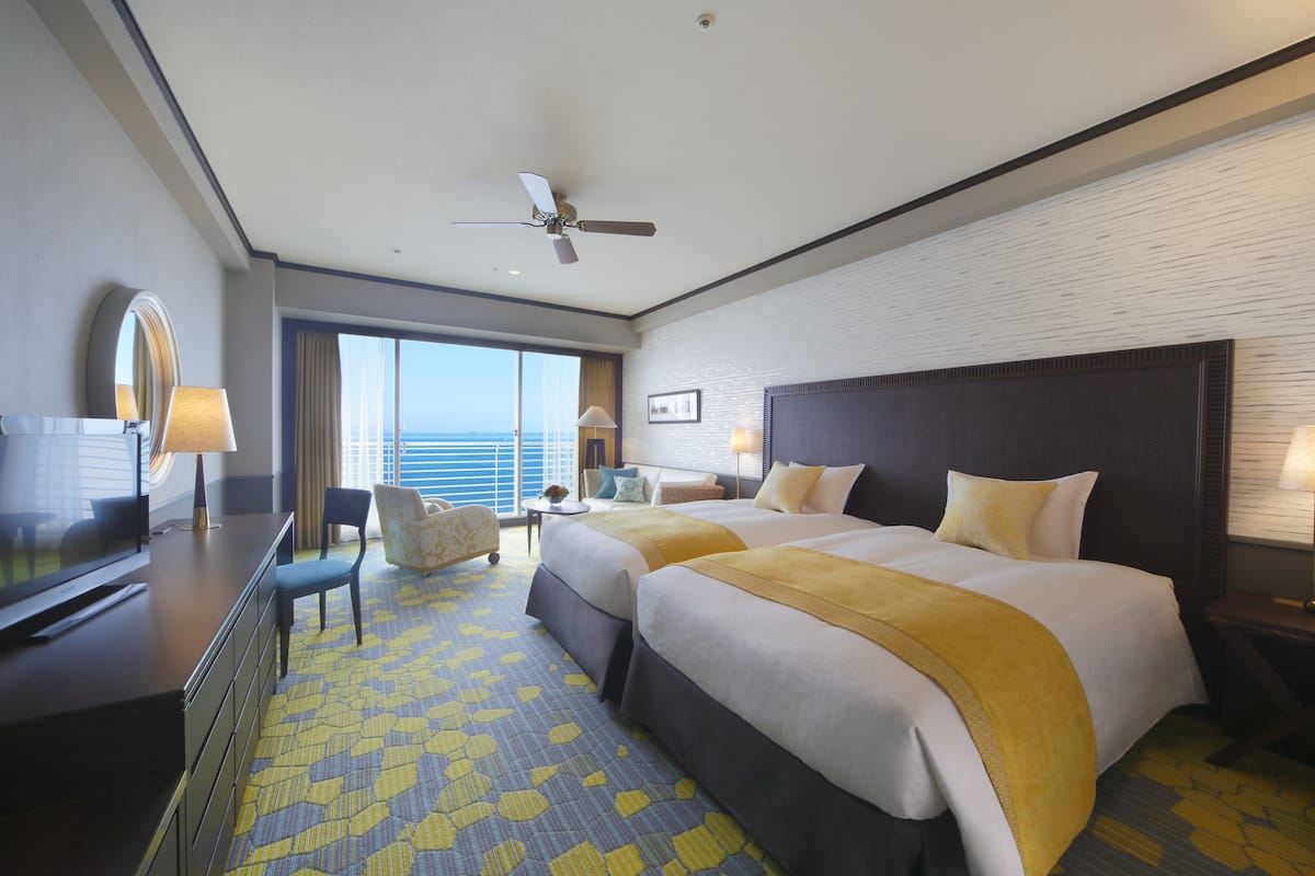 A hotel room with two beds, a desk with a TV, a chair, a small table with a lamp, and a view of the ocean.