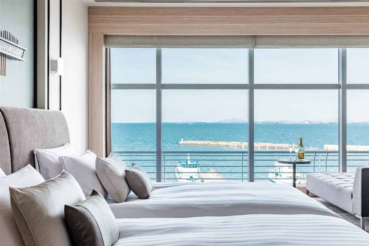 A hotel room with a large bed, a view of the sea, and a table with a bottle.