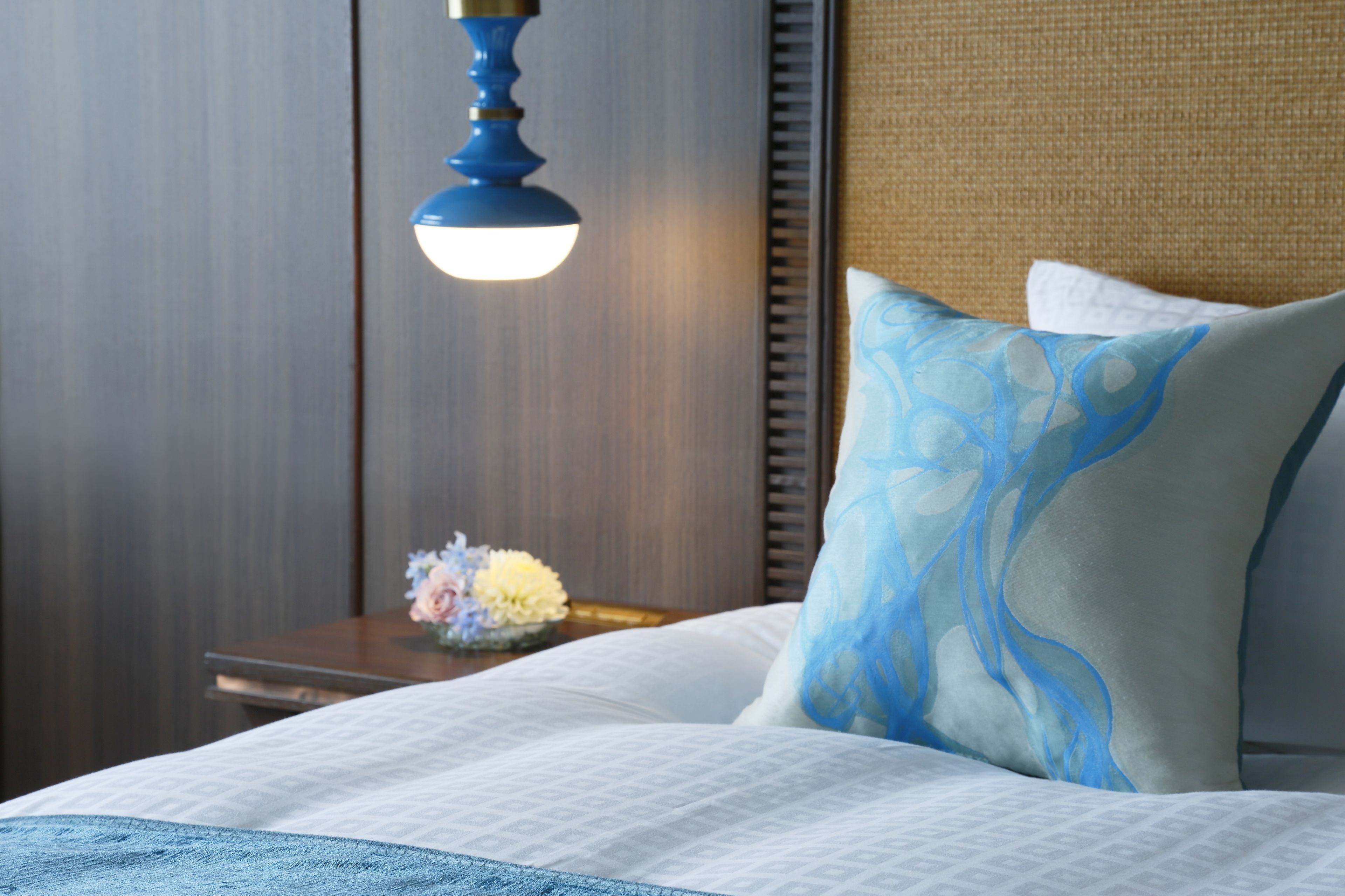 A neatly made bed with a patterned bedspread, a decorative pillow, a bedside table with a vase of flowers, and a wall-mounted lamp.