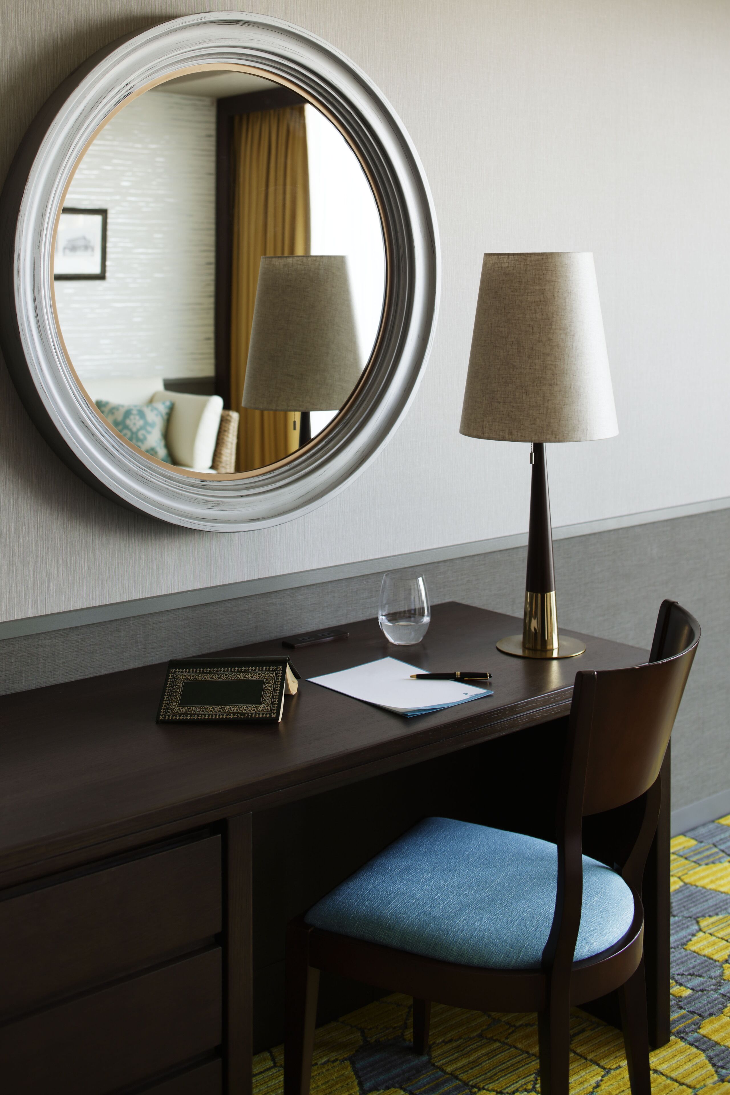 Mesa de quarto de hotel com um espelho, uma luminária, um copo de água, um caderno e uma caneta.