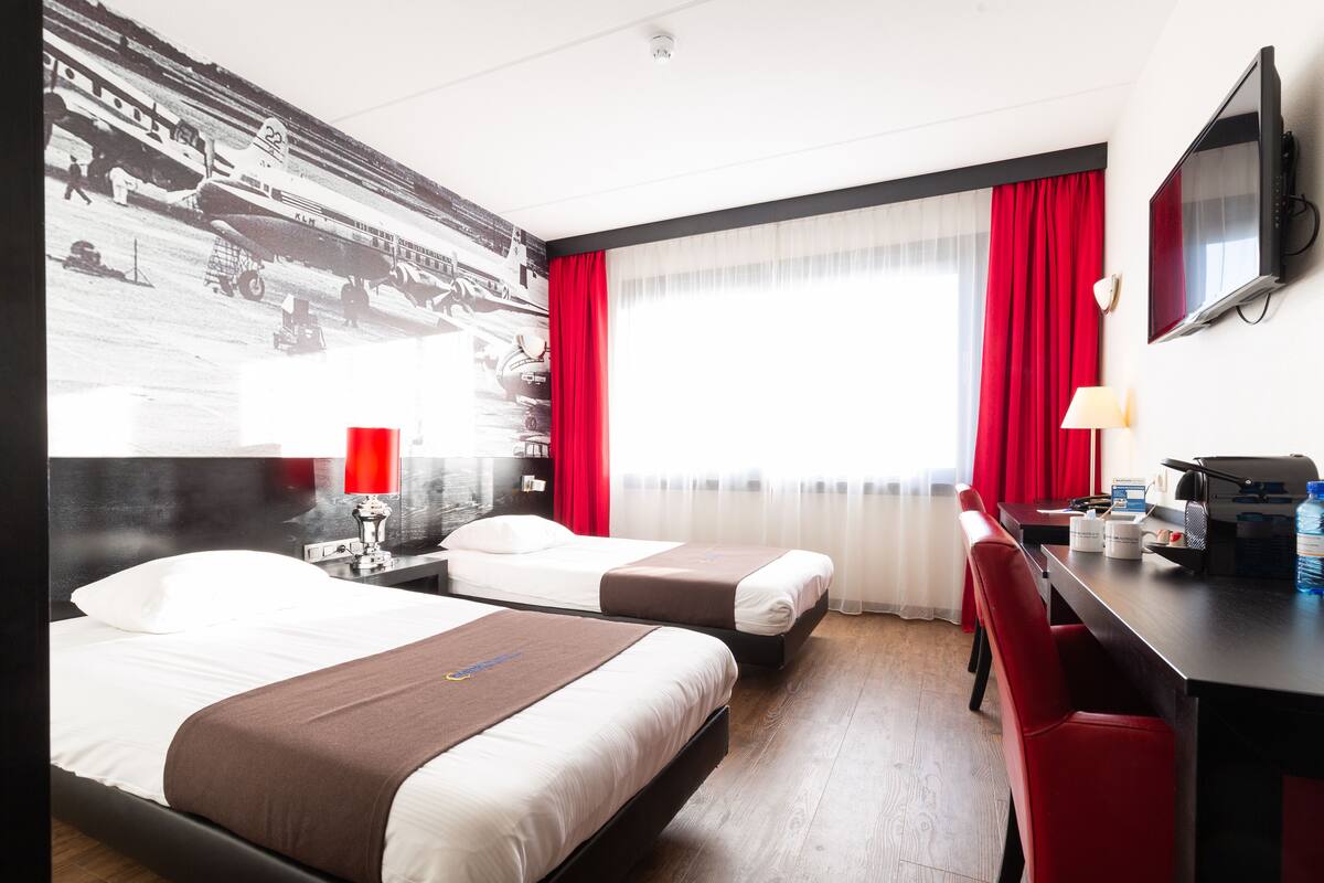 Habitación de hotel con dos camas, un escritorio, una silla, un televisor y un ventanal con cortinas rojas.