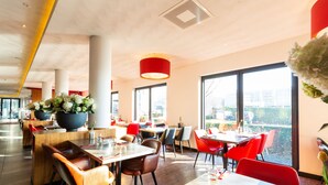 Un restaurante moderno con mesas y sillas de madera, ventanas amplias y una lámpara colgante roja.