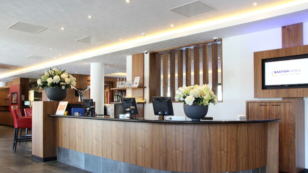 Un mostrador de recepción con detalles de madera, una planta en maceta grande y una pantalla digital que muestra el nombre del hotel.