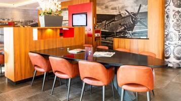 A modern hotel lobby with a wooden reception desk, a large round table with orange chairs, and a mural of an airplane on the wall.