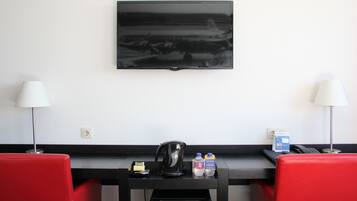 Ein Hotelzimmer mit einem an der Wand befestigten Flachbildfernseher, einem Schreibtisch mit Wasserkocher, zwei roten Stühlen, einer Kaffeemaschine und einem Mülleimer.