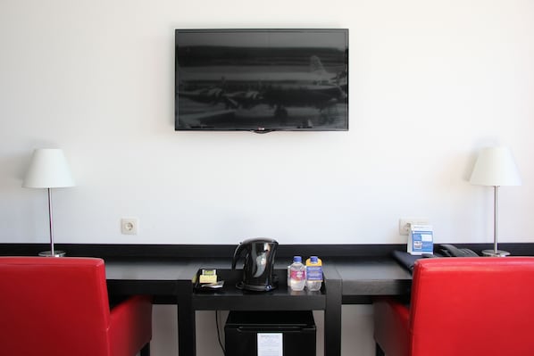 A hotel room with a flat-screen TV mounted on the wall, a desk with a kettle, two red chairs, a coffee machine, and a trash bin.