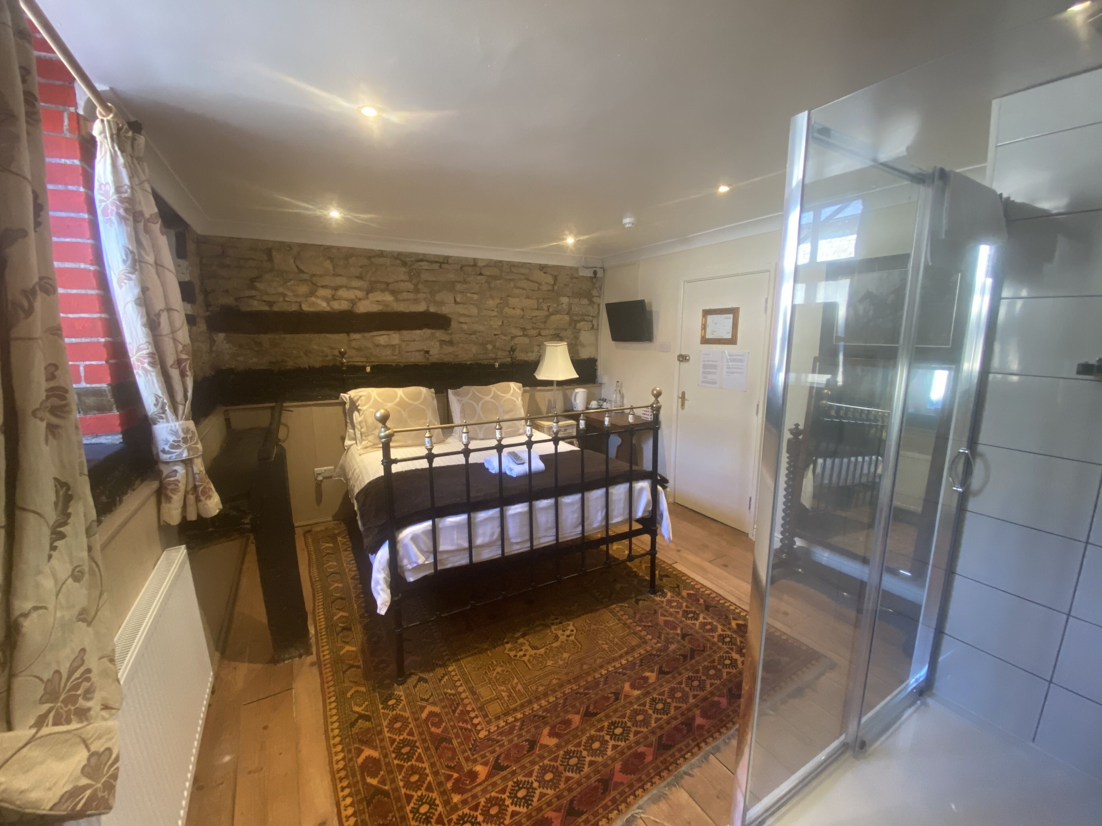 A bedroom with a bed, a nightstand, a lamp, a stone wall, a shower area, and a patterned rug.