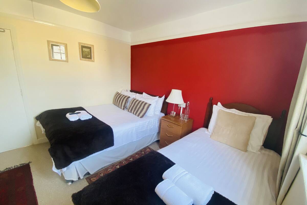 A hotel room with two beds, a red accent wall, a nightstand with a lamp, and a small window.